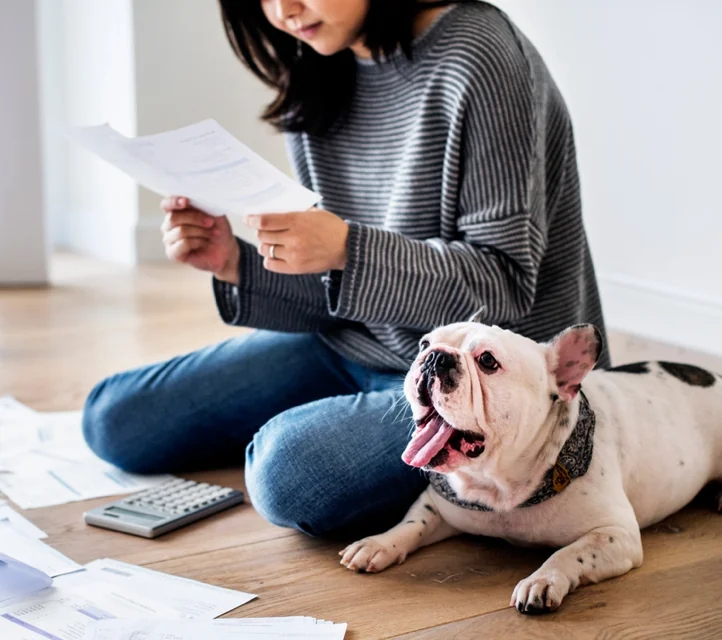 A woman doing taxes on a bare floor with a French bulldog looking bored beside her.