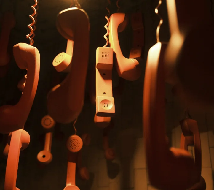 Many late 20th Century telephone handsets dangling by their cords in a dimly lit room.