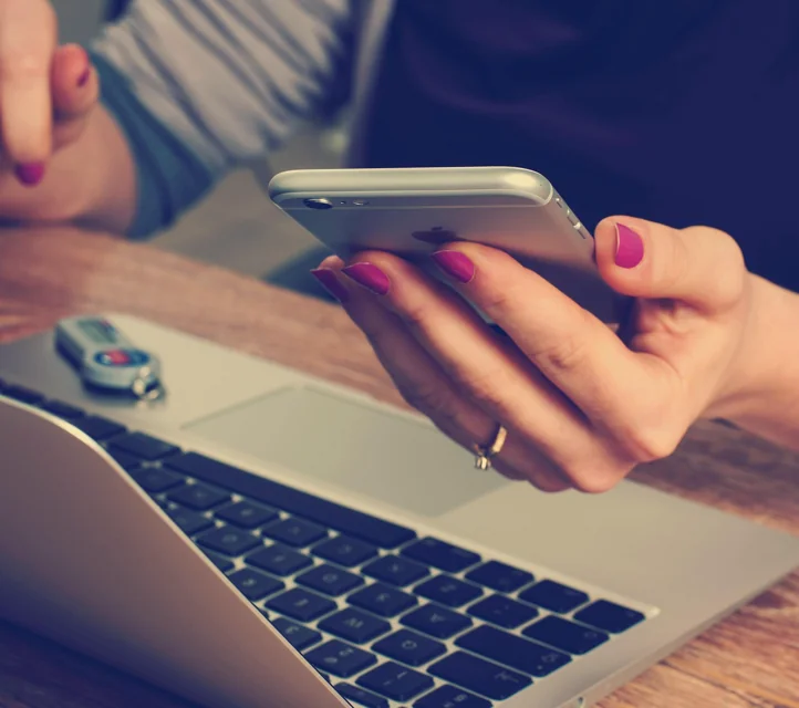 A hand holding a phone next to an open laptop as if both are required for the task depicted.