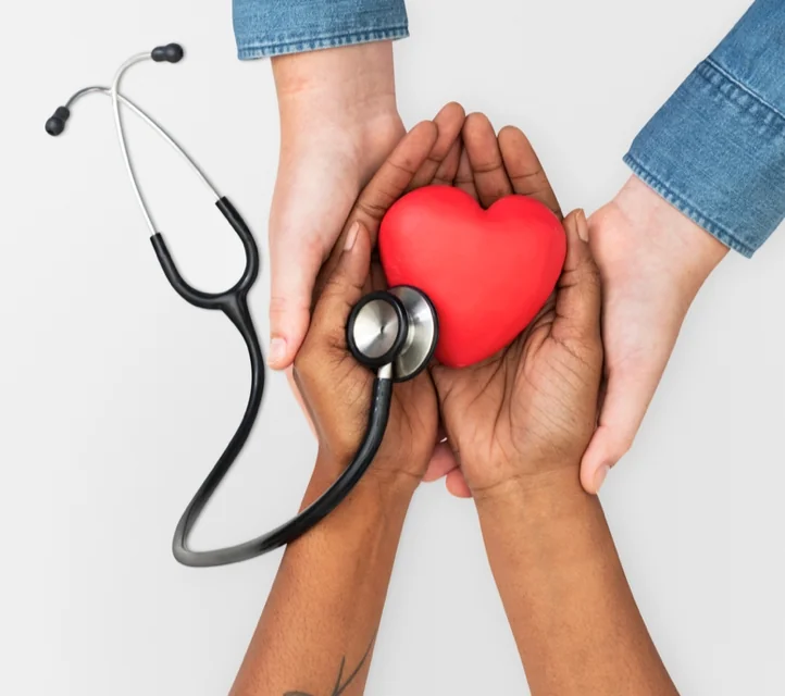 Two sets of hands cradling a heart with the diaphragm of a stethoscope resting on it.